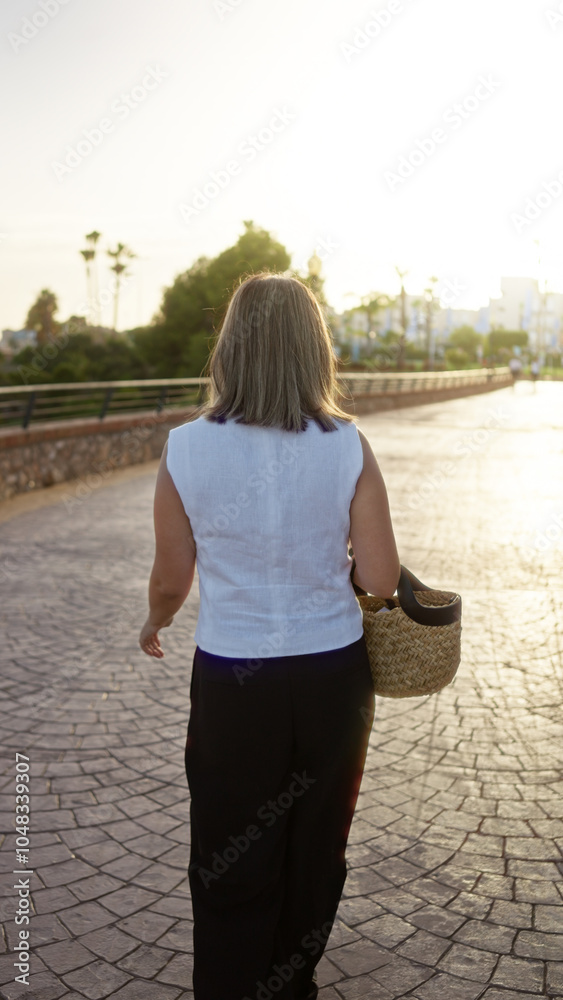 Woman walking outdoors at sunset carrying basket in sunny lush park with paved pathway and some trees and buildings visible in the background.