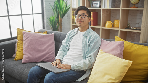 A young asian man in casual attire sits comfortably with a laptop on a couch in a modern living room.