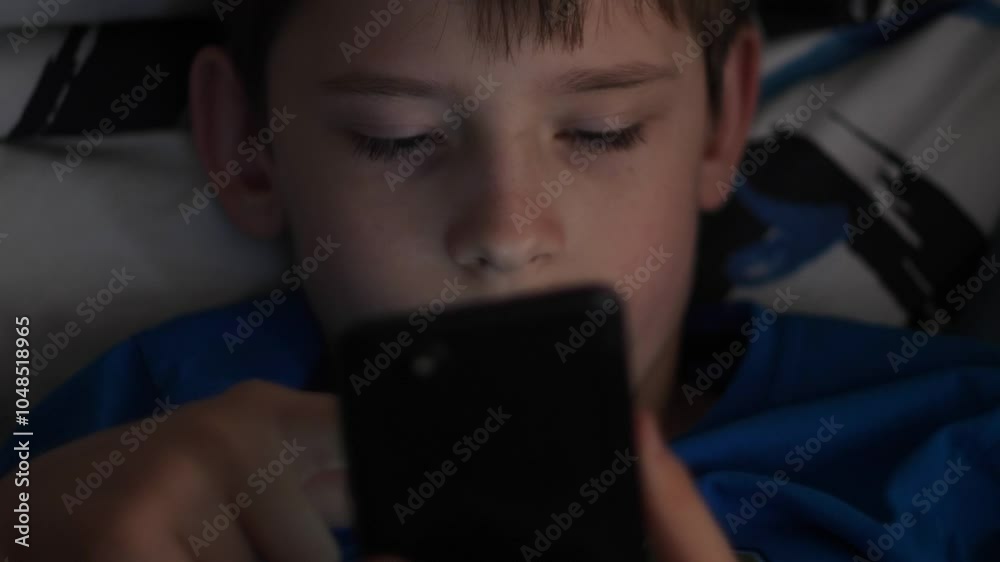 Caucasian boy 10 years old enthusiastically uses smartphone in the dark scrolls on the screen, teenager uses gadget lying in bed in the dark