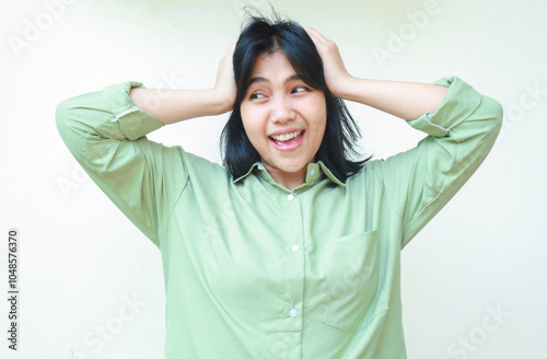 stressed asian woman with dark hair smiling crazy and grabbing her head looking away wearing green oversized shirt, frustrated female feel under pressure, isoated on white background photo