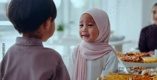 During Eid al-Fitr, a young Muslim boy hugs his sister to apologize, with a table full of snacks and food in the background. Relatives and neighbors visit, celebrating forgiveness and bonding photo