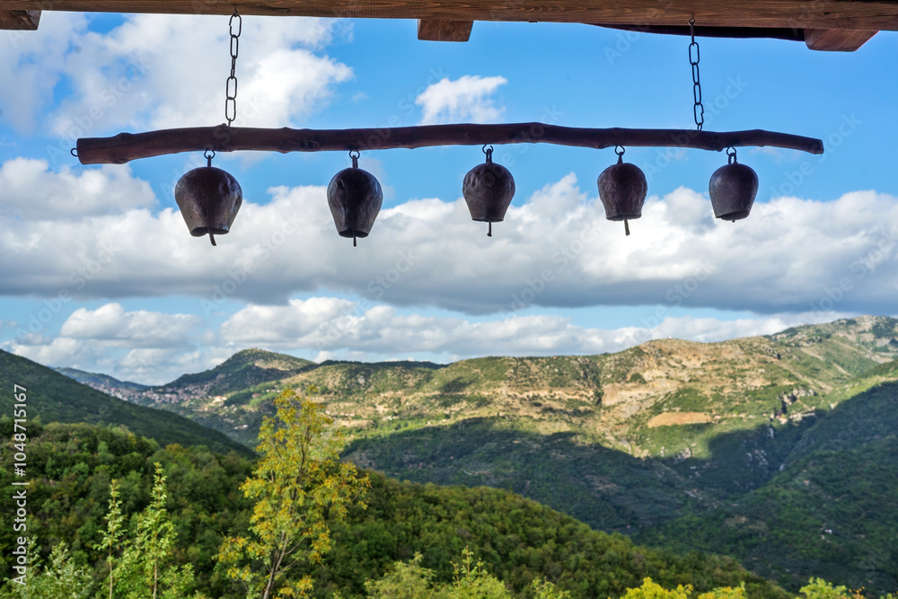 Traditional metal bells hanging from a wooden bar, framing a view of the lush, mountainous landscape in Zerzova, Arkadia, Greece.