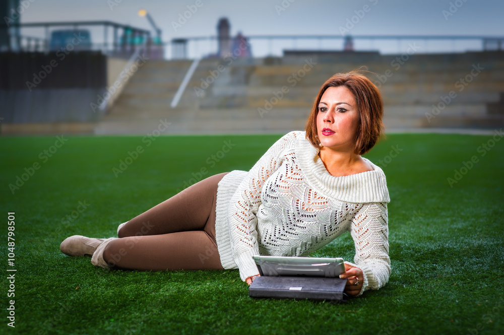 A woman in a white sweater and brown pants is lounging on grass, holding a tablet. The background features a blurred cityscape, suggesting an urban park setting.
