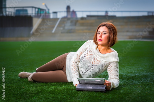 Obraz na płótnie A woman in a white sweater and brown pants is lounging on grass, holding a tablet. The background features a blurred cityscape, suggesting an urban park setting. w salonie
