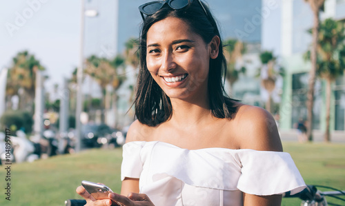 Half length portrait of toothy Latino female tourist holding modern digital technology in hand and smiling during vacations journey to Spain, joyful millennial blogger enjoying smartphone using #1048957117