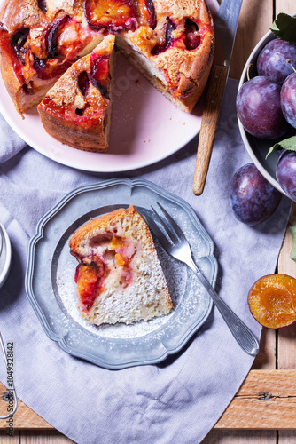 Plum pie with filling and sugar crust, sprinkled with powdered sugar and plums on a wooden table.