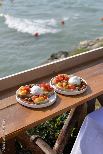 2 piatti con bruschette con pomodori, mozzarelle e olive servite su di un tavolo difronte al mare