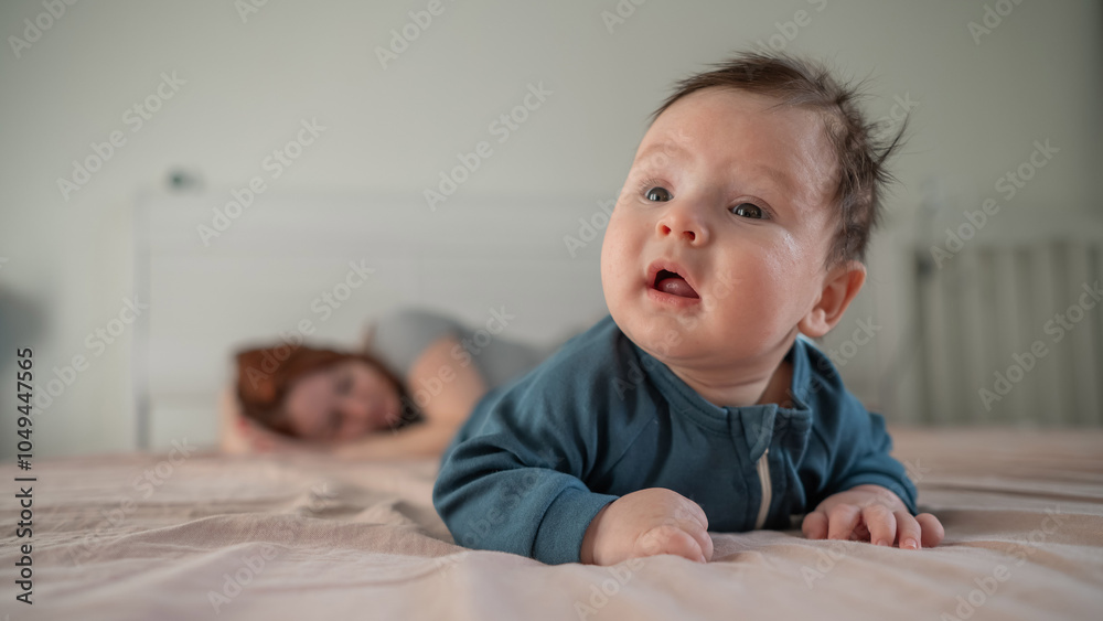 A three-month-old boy lies on his stomach on the bed and his mother sleeps behind him. Postpartum depression. A three-month-old boy lies on his stomach on the bed and his mother sleeps behind him. Postpartum depression.