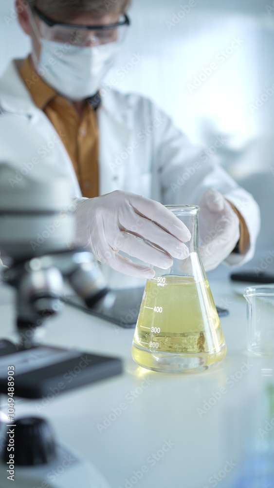 Fototapeta premium Man scientist researcher, wearing white protective gloves, mask and glasses, is holding a yellow chemical solution inside Erlenmeyer flask in a laboratory, vertical portrait view. Science and medicine