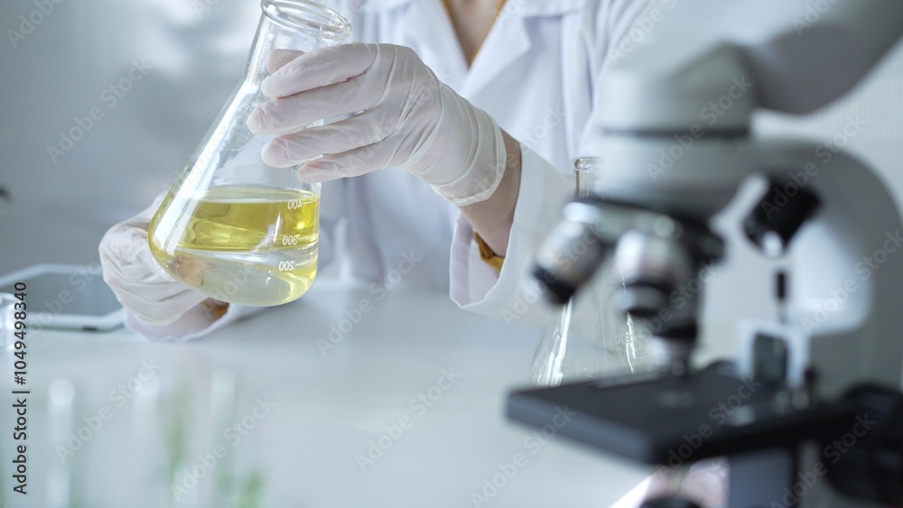 Microscope is near a scientist, wearing a lab coat and white protective gloves, and holding beaker with a yellow oily liquid in laboratory setting, close up. Medicine and science concept Microscope is near a scientist, wearing a lab coat and white protective gloves, and holding beaker with a yellow oily liquid in laboratory setting, close up. Medicine and science concept