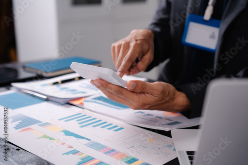 businessman sits at his office desk, analyzing financial documents and graphs on his laptop. paperwork, calculator, and charts, he strategizes investments, audits, tax planning for success.