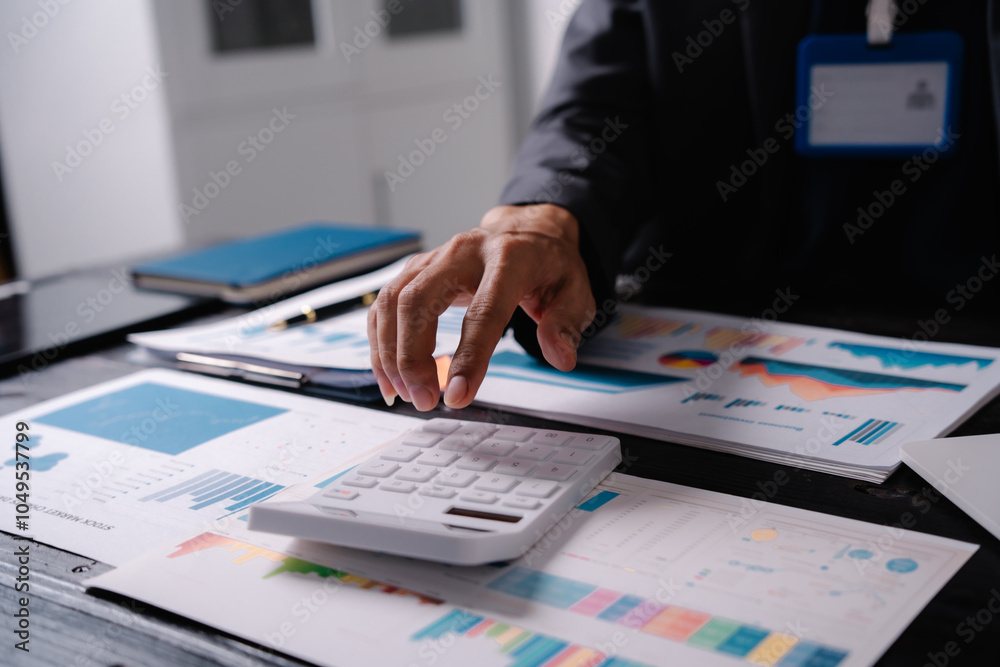 businessman sits at his office desk, analyzing financial documents and graphs on his laptop. paperwork, calculator, and charts, he strategizes investments, audits, tax planning for success.
