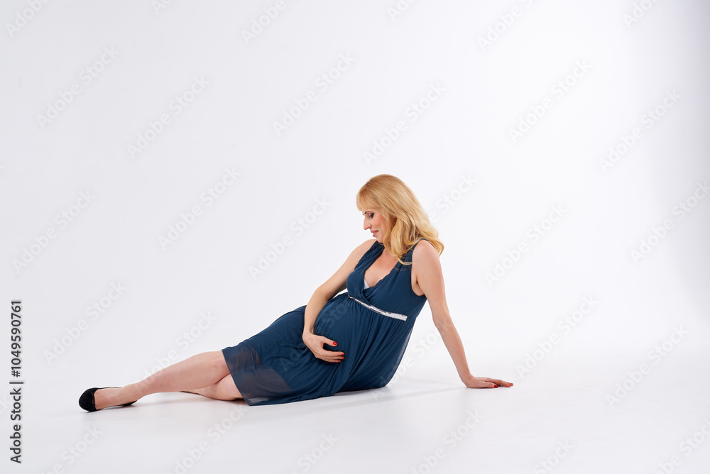 A pregnant woman is wearing a blue dress and standing in front of a white background. She is wearing black heels and has blonde hair A pregnant woman is wearing a blue dress and standing in front of a white background. She is wearing black heels and has blonde hair