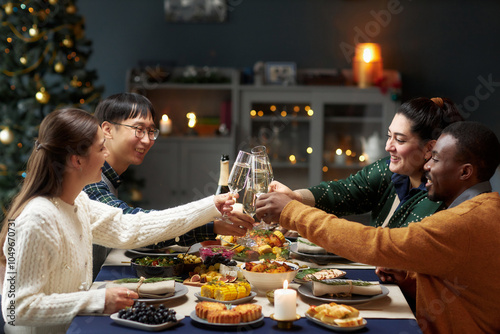 Side view of diverse group of friends clinking glasses sitting at dining table in living room while celebrating Christmas together at home, copy space photo