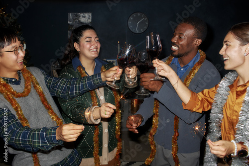 Medium shot of diverse group of cheerful friends wearing vintage outfits and tinsels clinking glasses with wine while celebrating Christmas together in living room at home, camera flash photo