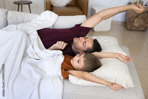 Calm sleepy awaking Latin dad and son stretching bodies with closed eyes, lying under white blanket, resting in beddings on home couch close together, waking up in morning after sleeping