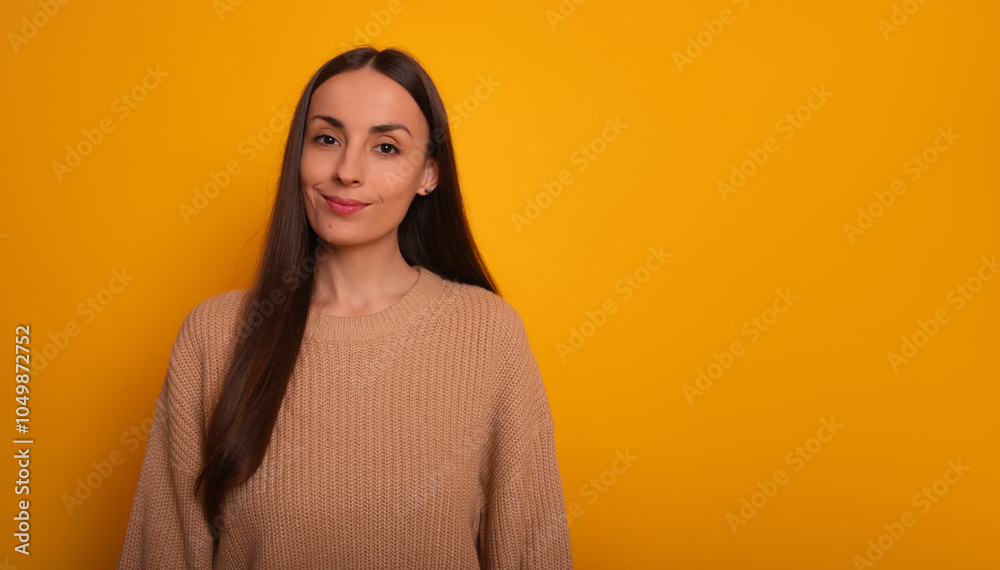 Fototapeta premium A young woman with long brown hair stands against a bright yellow background, wearing a beige sweater and smiling slightly. The image is cheerful and friendly, conveying warmth and positivity.