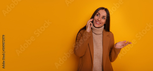 A cheerful woman in a brown coat talks on her smartphone against a bright yellow background, gesturing with her hand. Ideal for communication, business, or lifestyle themes. photo