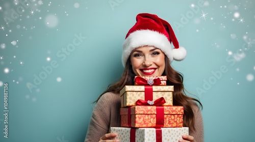 Smiling woman in Santa hat holding a stack of Christmas presents against a teal snowy background. Festive cheer!