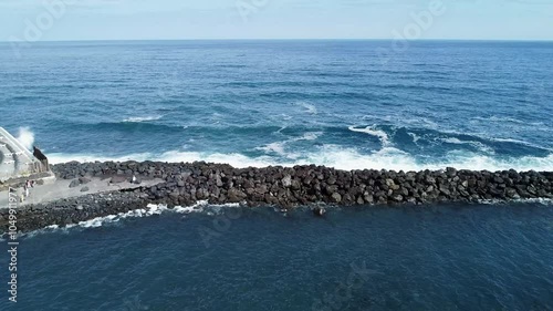 Aerial view of ocean waves crashing against a rugged stone breakwater, creating contrast between the deep blue sea and the coastal barrier