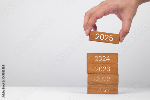 A hand placing a wooden block labeled '2025' on top of stacked blocks representing previous years. Symbolizes progress and planning.