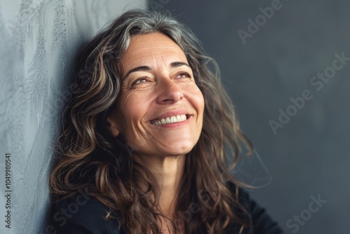 A woman with gray streaked hair leans against a wall, gazing upwards with a content expression, capturing a moment of reflection and contemplation indoors. photo