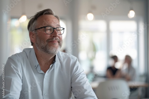 Portrait of a mature man wearing glasses in a cozy office setting, thoughtfully looking out the window with a gentle smile, conveying optimism and confidence.