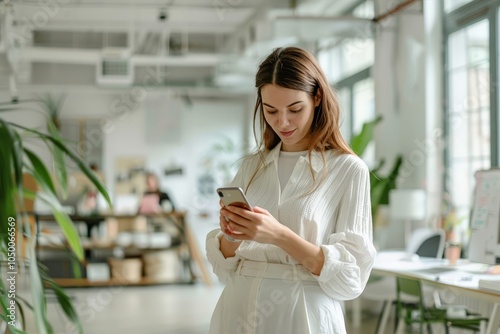 A professional woman in a white blouse is engrossed in her phone within a spacious, plant-filled office environment, symbolizing connectivity and modern work ethics.