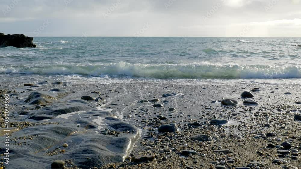 A rocky shore with small, smooth stones and larger rocks. White foam from the waves is receding, leaving streaks of water on the rocks. A rocky shore with small, smooth stones and larger rocks. White foam from the waves is receding, leaving streaks of water on the rocks.