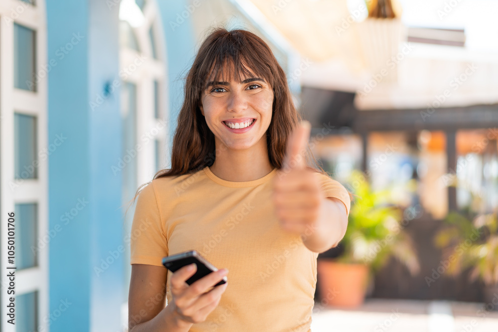Young woman at outdoors using mobile phone while doing thumbs up Young woman at outdoors using mobile phone while doing thumbs up