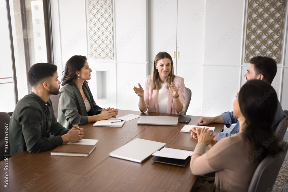 Fototapeta premium Positive young project leader woman holding meeting with team, talking to employees, coworkers at large table, offering marketing strategy, teamwork plan, ideas for brainstorming