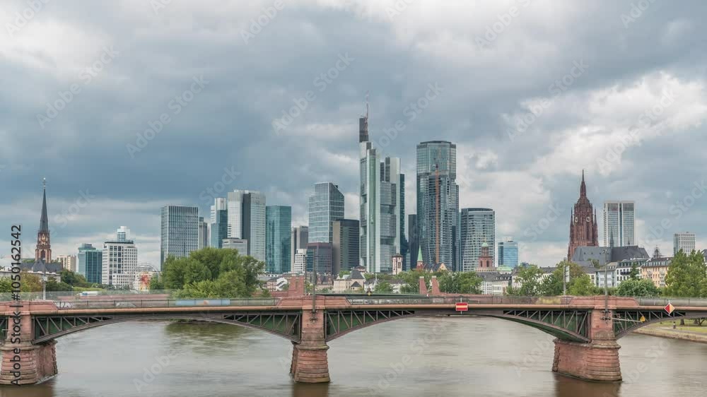 Aerial hyperlapse of Frankfurt skyline with Main River and Ignatz Bubis Bridge. Skyscrapers and church spires reflected in the water beneath dramatic clouds timelapse. Frankfurt, Germany