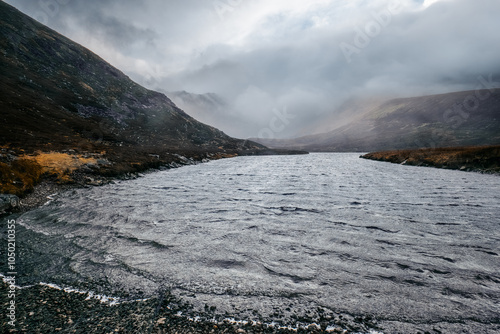 A misty, brooding landscape with rolling hills and a winding, gray river at its center, creating a scene of quiet solitude and introspective calm. photo