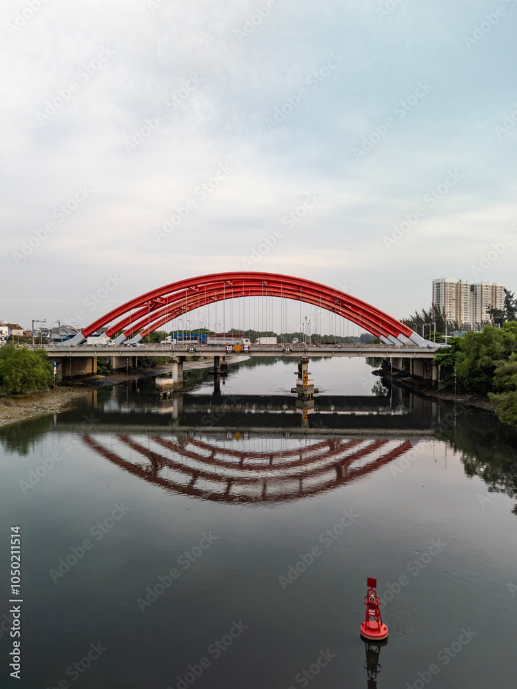 Fototapeta premium Traffic bridge over river in Ho Chi Minh City, Vietnam from aerial view featuring two large red steel arches and a wide, ten lane highway for heavy vehicles. Low angle with reflection on water