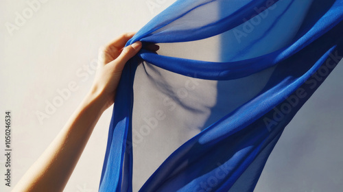 A hand gracefully holds a transparent blue piece of fabric cast with intriguing shadows, creating a play between light and dark and inviting a thoughtful reflection.