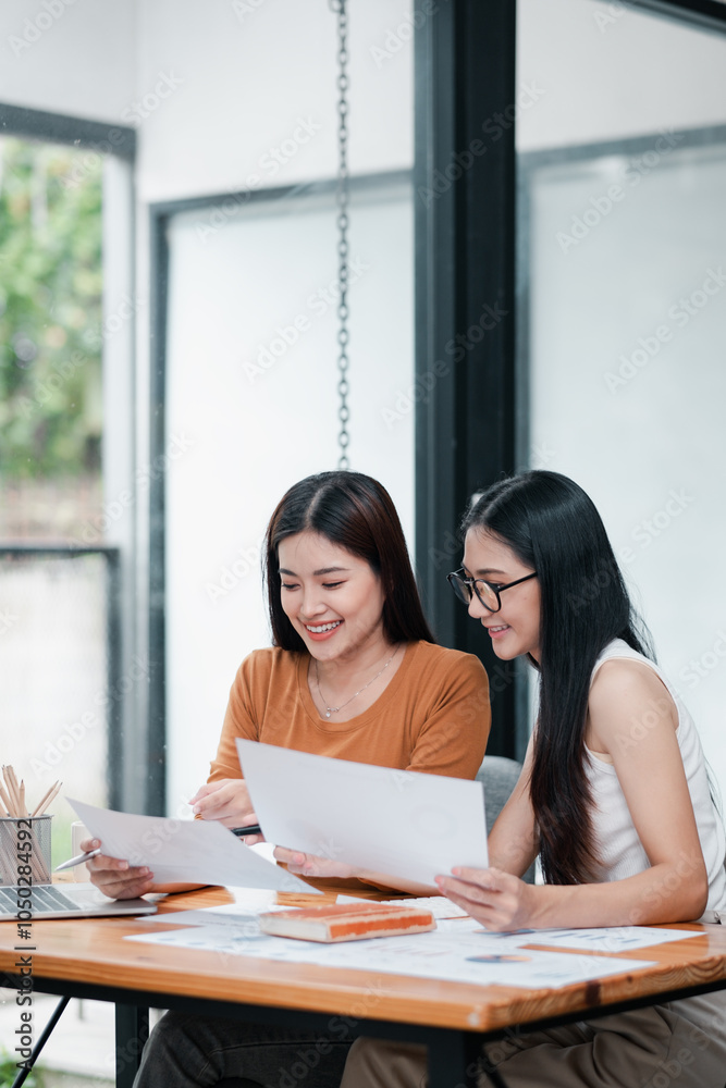 Two young women working together at a desk, reviewing documents in a bright, modern office setting.