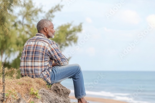 An African grandfather of 65 years enjoys a moment of tranquility while looking out over the ocean horizon from a cliff. The soft dawn light creates a peaceful atmosphere, enhancing the minimalist bac