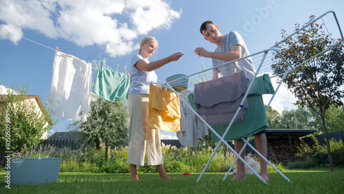 Schilderij op canvas A woman and a man hanging clothes on a drying rack and clothesline in a bright backyard, enjoying a sunny day doing household chores together