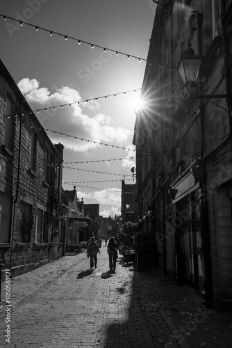 A black and white silhouette of two people walking up a quiet, cobbled street with strings of lights above them, Harrogate, Yorkshire, UK.