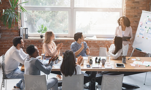 Black creative lady speeking in front of young business team in office, colleagues cheering woman with applause photo