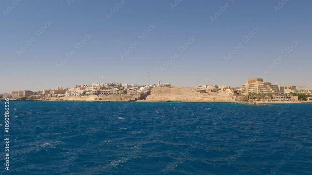 A panoramic view of the skyline of Hurghada, Egypt, nestled on sandy hills. A yacht sails in the sea near the city. A panoramic view of the skyline of Hurghada, Egypt, nestled on sandy hills. A yacht sails in the sea near the city.