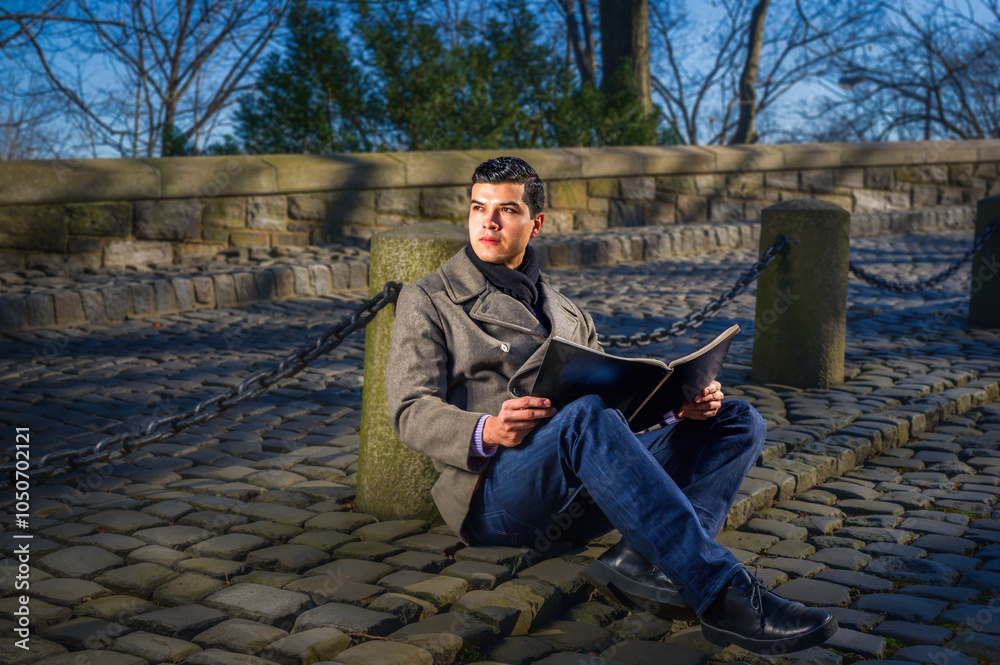 A man sits on a cobblestone path, reading a book and enjoying a sunny day. The setting is serene, with a stone wall and leafless trees in the background.