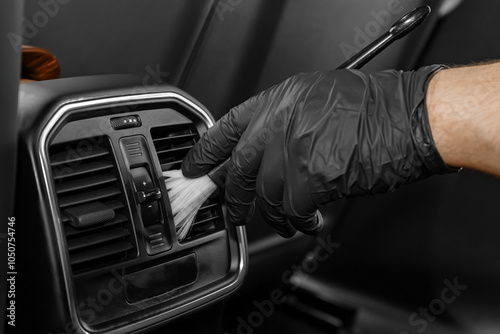 A man in black rubber gloves cleans dust from air ducts inside a car with a brush. Detailing studio photo