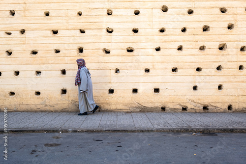 Composition of an elderly woman walking down the street dressed in a grey djellaba in front of the massive city wall of Fez in Morocco, North Africa photo