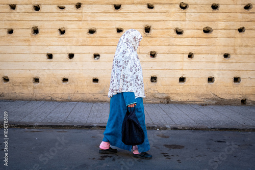 Composition of an elderly woman walking down the street dressed in a blue djellaba and a hijab in front of the massive city wall of Fez in Morocco, North Africa photo