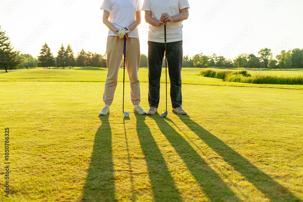 Fototapeta premium man and woman playing golf on the course at sunset, close-up of golf ball and clubs and people's feet on green grass, couple competing in the game and hitting the ball into the hole
