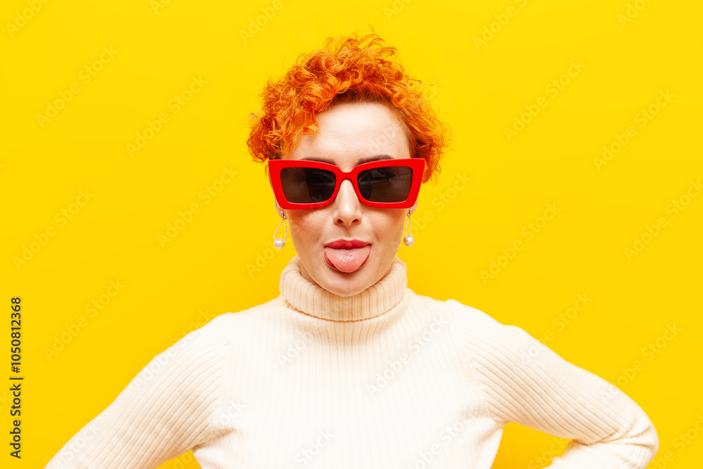 young red-haired woman with curly hair in sunglasses shows her tongue on a yellow isolated background, girl with beautiful make-up in red glasses