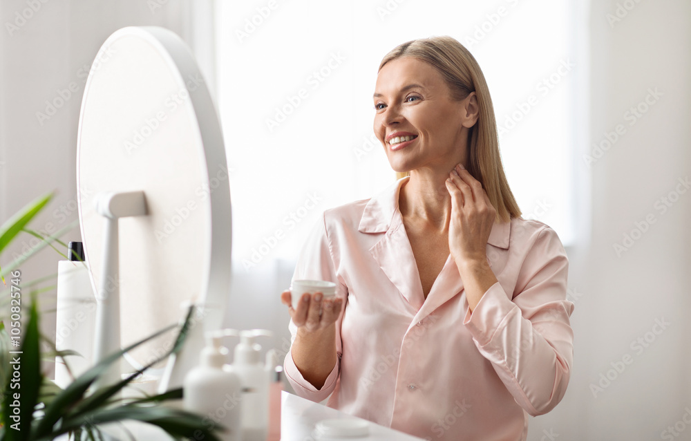 Beautiful middle aged woman applying moisturizing cream on her neck while sitting near mirror at home, attractive mature female enjoying domestic beauty routine, holding jar with nourishing lotion