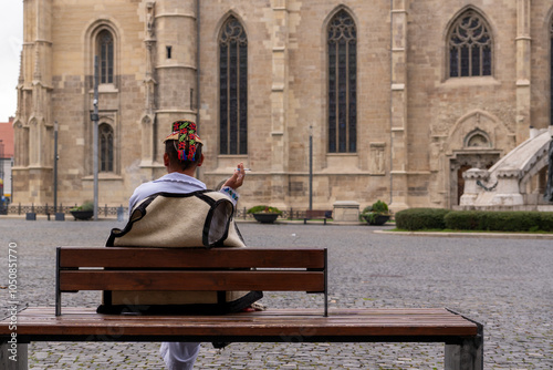 Cluj-Napoca, Romania, October 20, 2024: A man dressed in traditional Romanian clothing sits on a bench in the central square, smoking near St. Michael's Church.