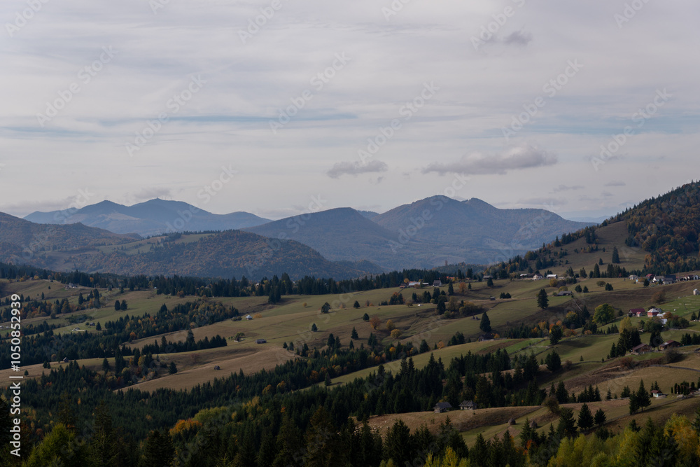 A scenic view of the Carpathian Mountains with rolling hills, forests, and scattered houses under a cloudy sky. Early autumn colors are visible in the landscape. A scenic view of the Carpathian Mountains with rolling hills, forests, and scattered houses under a cloudy sky. Early autumn colors are visible in the landscape.